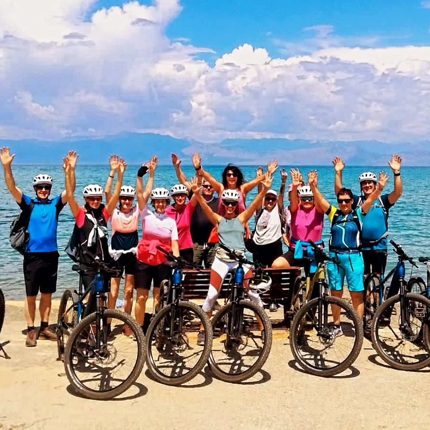 Eine Gruppe von zwölf Menschen mit E-Bikes am Strand, alle reißen die Arme hoch
